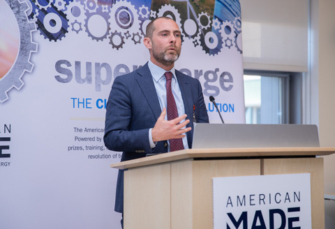A man wearing a suit speaks from behind a podioum with a sign that says, “American Made.”