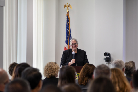 A woman stands at the front of a room with a microphone, the audience in the room looks at her