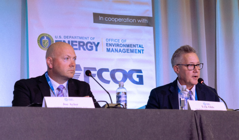 Two individuals sitting at a table on a stage speaking on a panel at a conference