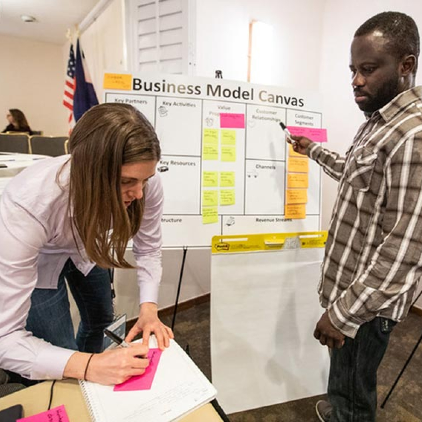 Image of a female writing on writing on a pink sticky note as a male colleague standing next to a white board looks on.