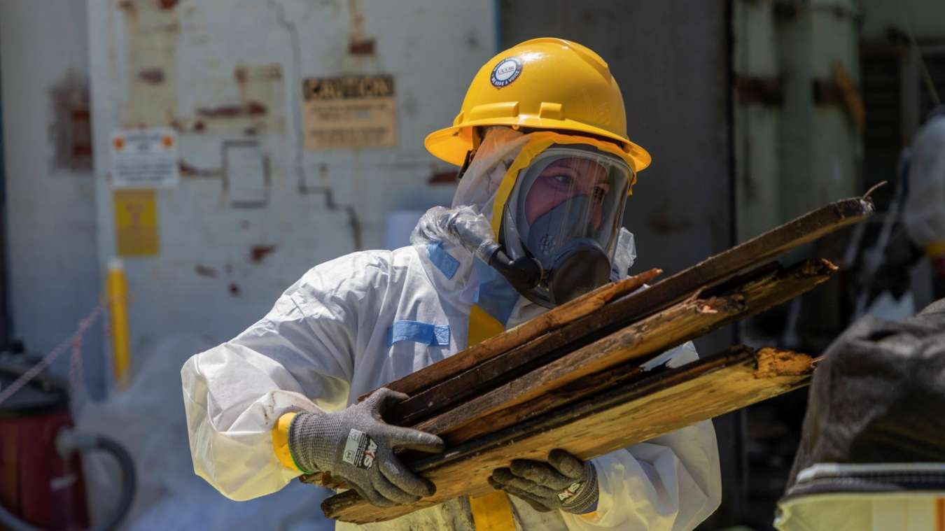 A person in protective gear carries wood in their arms.