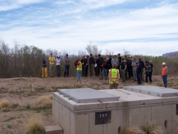 As Durham University students stand on the bank of the Colorado River, Moab Groundwater Manager Ken Pill (in yellow vest at left) explains how the groundwater interim action system operates. A well vault is in the foreground. As Durham University students stand on the bank of the Colorado River, Moab Groundwater Manager Ken Pill (in yellow vest at left) explains how the groundwater interim action system operates. A well vault is in the foreground.