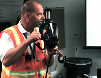 Advanced Mixed Waste Treatment Project Industrial Hygiene Manager Roger Raymond demonstrates breathing equipment. The specially-designed breathing equipment includes a primary face-piece and a second stage face-piece regulator, which is attached to a manifold configured with dual air supply (main and secondary), a vortex cooler, and an escape bottle. This configuration allows workers to safely enter atmospheres in excess of 100,000 derived air concentration levels. Advanced Mixed Waste Treatment Project Industrial Hygiene Manager Roger Raymond demonstrates breathing equipment. The specially-designed breathing equipment includes a primary face-piece and a second stage face-piece regulator, which is attached to a manifold configured with dual air supply (main and secondary), a vortex cooler, and an escape bottle. This configuration allows workers to safely enter atmospheres in excess of 100,000 derived air concentration levels.