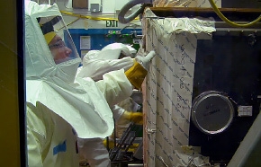 Workers pull a glove box apart and encase each section in a protective covering to prevent exposure. Workers pull a glove box apart and encase each section in a protective covering to prevent exposure.
